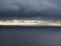 Dark grey clouds over the sea, outline of coastline in the distance.