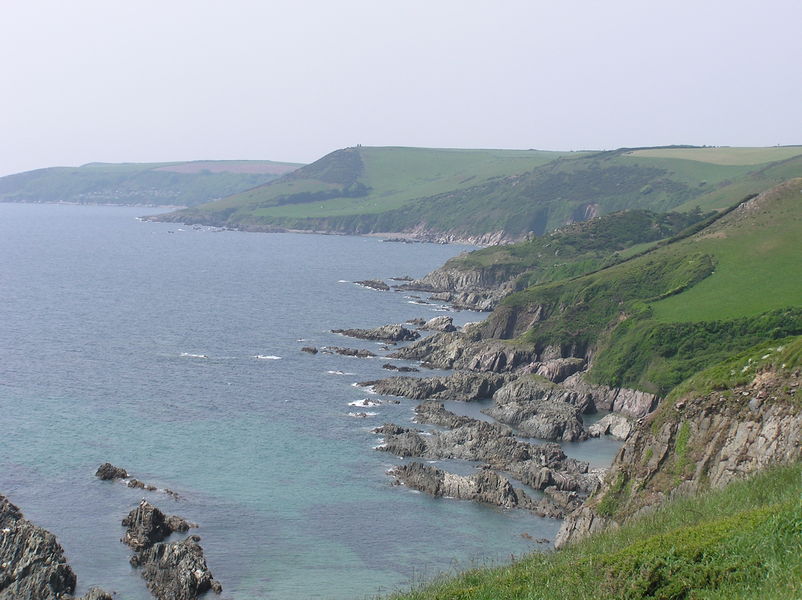 Looking along coastline, grey rocks at shoreline, green fields above.