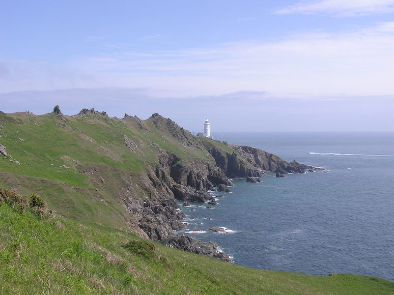 Sun covered sharp ridge leading down to white lighthouse, steep rocky cliffs and inlets beside the sea.