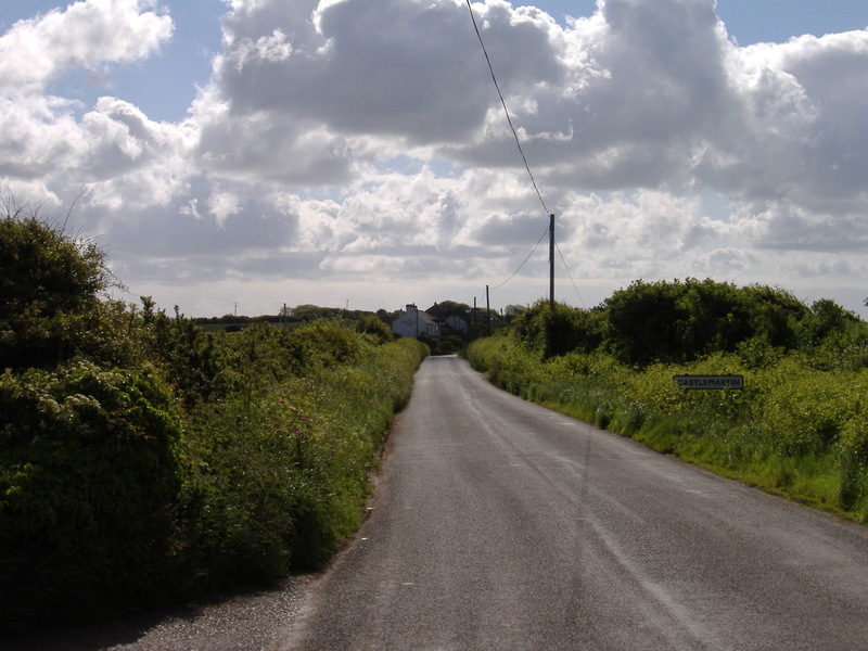 View down road with grassy sides, houses just visible in distance, sunny but lots of clouds above
